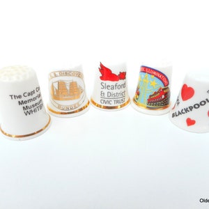 May include: Five white ceramic thimbles with gold trim and colorful designs. The thimbles feature images of a ship, a red bird, a Ferris wheel, and hearts with the word "Blackpool".