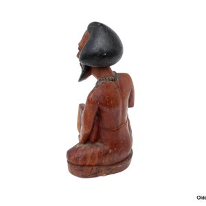May include: A wooden figurine of a person with a black turban, sitting with their legs crossed. The figurine is carved from a reddish-brown wood and has a detailed face.