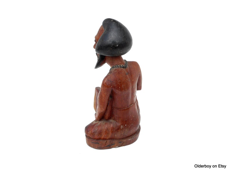 May include: A wooden figurine of a person with a black turban, sitting with their legs crossed. The figurine is carved from a reddish-brown wood and has a detailed face.