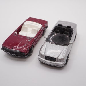 May include: Two toy convertible cars, one red and one silver, with black interiors. The red car has a white interior and the silver car has a black interior.