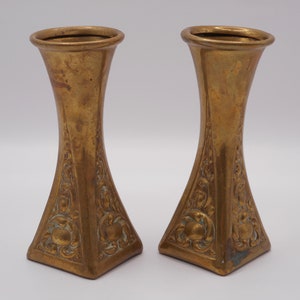 May include: Two small, ornate brass vases with a square base and a flared top. The vases have an intricate, floral design.
