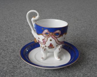 May include: A white porcelain teacup with a blue and gold rim and a pink ribbon design. The cup has three legs and a handle shaped like a swan's neck. The cup is sitting on a matching saucer.