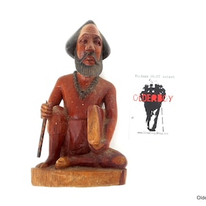 May include: Hand-carved wooden figurine of a seated man with a beard and a hat. The figurine is painted in shades of brown and red. The man is holding a staff in his right hand and a shield in his left hand. The figurine is sitting on a wooden base.