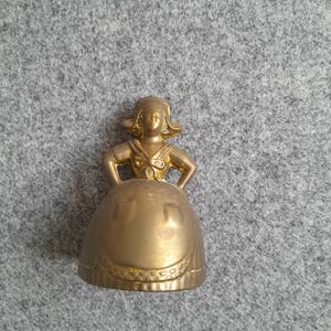 May include: A metallic gold-toned bell shaped like a figure wearing a traditional bonnet, a bodice with a bow at the neckline, and a wide skirt featuring a prominent apron design. The bell has a smooth, reflective surface and a flared base.