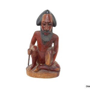 May include: A wooden figurine of a seated man with a black hat and beard. The figurine is carved in a reddish-brown color and has a detailed face.