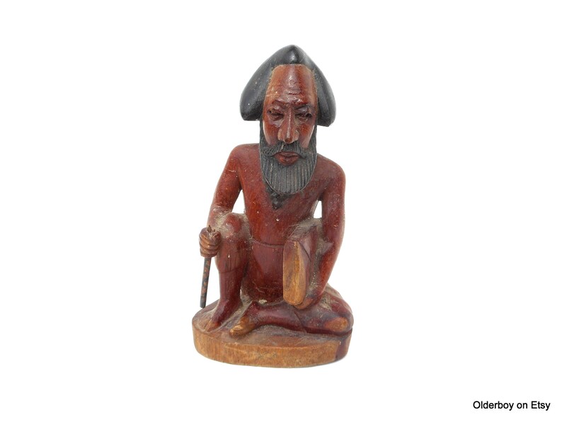 May include: A wooden figurine of a seated man with a black hat and beard. The figurine is carved in a reddish-brown color and has a detailed face.