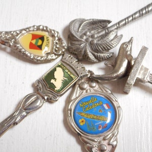 Könnte beinhalten: Eine Sammlung von Vintage-Souvenirlöffeln mit dekorativen Griffen. Die Löffel weisen verschiedene Designs auf, darunter einen Delfin, eine Palme und Embleme mit Flaggen und Text. Die Löffel sind aus silberfarbenem Metall gefertigt und haben farbige Emaille-Details.