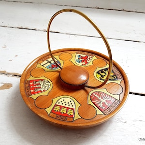 Könnte beinhalten: Ein Holztablett mit einem goldenen Griff. Das Tablett ist mit sechs bunten Wappen verziert, jedes mit einem anderen Städtenamen:  "Hamburg", "Köln", "Nürnberg", "München", "Frankfurt" und "Bonn".