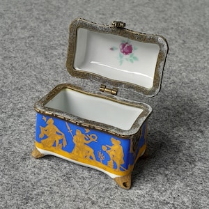 Könnte beinhalten: Geöffnete, rechteckige Schmuckschatulle mit Klappdeckel. Die Schatulle ist blau mit goldenen Figuren und Verzierungen. Der Innenraum ist weiß mit einem rosa Rosenmuster. Die Schatulle hat kleine, goldfarbene Füße.
