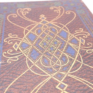 K&ouml;nnte beinhalten: Ein brauner Buchumschlag mit einem komplizierten goldenen und blauen Design. Das Design zeigt ein sich wiederholendes Muster aus Knoten und Wirbeln.