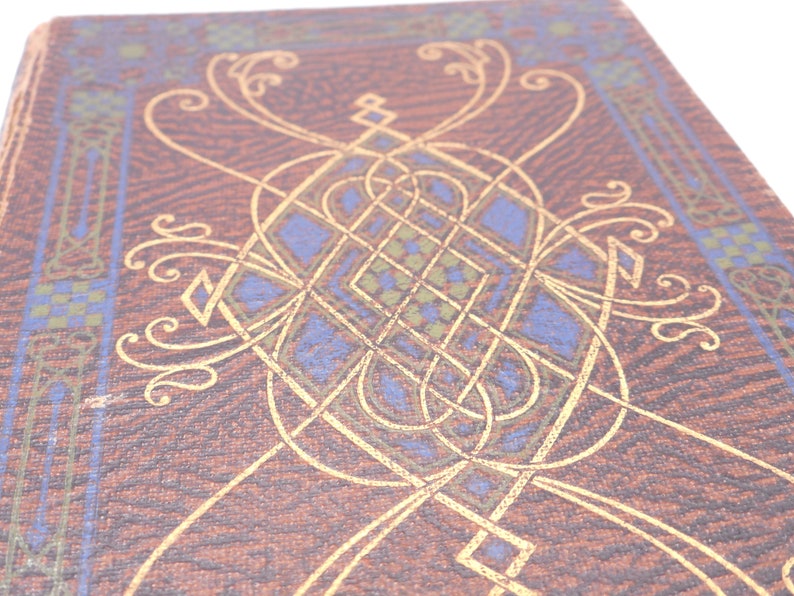 K&ouml;nnte beinhalten: Ein brauner Buchumschlag mit einem komplizierten goldenen und blauen Design. Das Design zeigt ein sich wiederholendes Muster aus Knoten und Wirbeln.