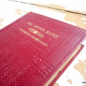 Pode incluir: Um livro encadernado em vermelho com letras douradas na capa. O título é "The Diva's Ruby" de F. Marion Crawford.