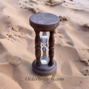 以下が含まれることがあります： 砂の背景に置かれた、濃い茶色の木製砂時計。砂時計は、白い砂で満たされた透明なガラスの中心部を備えています。ベースと上部は円形で、デザインには装飾的な旋盤加工された脚が含まれています。「Olderboy.etsy.com」のテキストが表示されます。
