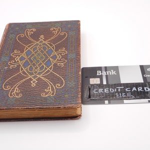 K&ouml;nnte beinhalten: Ein Vintage-Buch mit einem braunen Lederumschlag und einem goldenen und blauen Dekormuster. Das Buch liegt neben einer Kreditkarte mit dem Text "CREDIT CARD SIZE" darauf.
