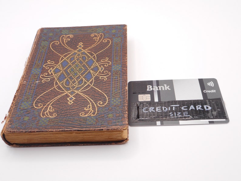 K&ouml;nnte beinhalten: Ein Vintage-Buch mit einem braunen Lederumschlag und einem goldenen und blauen Dekormuster. Das Buch liegt neben einer Kreditkarte mit dem Text "CREDIT CARD SIZE" darauf.
