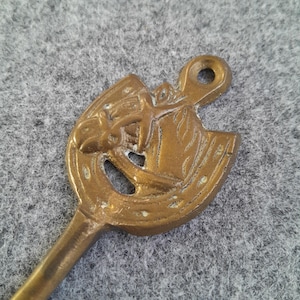 May include: A vintage brass key with a horseshoe-shaped head. The head features a detailed relief of a horse and rider. The key has a loop at the top for hanging and a long, slender stem.