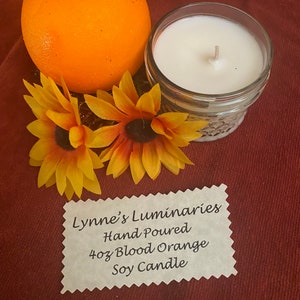 May include: A 4 oz soy candle in a clear glass jar with a white wax candle and a wick. The candle is labeled "Lynne's Luminaries Hand Poured 4 oz Blood Orange Soy Candle".