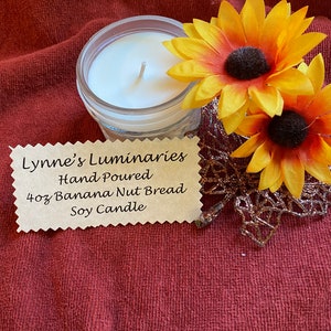 May include: A 4 oz soy candle in a clear glass jar with a white wax top. The candle is labeled "Lynne's Luminaries Hand Poured 4oz Banana Nut Bread Soy Candle". The candle is surrounded by two yellow artificial sunflowers.