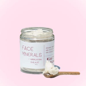 May include: A glass jar of face mineral cream with the text "FACE MINERALS + HIMALAYAN SHILAJIT 6oz" on the label. The cream is a light beige colour and is scooped out on a wooden spoon. The background is a light pink.