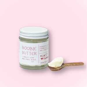以下が含まれることがあります： ピンクのラベルが付いた白い瓶に「Boobie Butter」と書いてあり、その下に「Violet Leaf and Poke Root」と「2oz」の文字が書かれている。瓶はピンクの表面の上に置かれている。小さな木のスプーンに白いバターが乗っているのが手前にある。