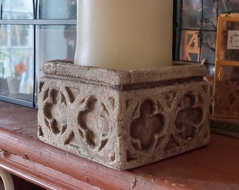 May include: A decorative candle holder made of stone with a carved design. The holder is square and has a light brown color. A white candle sits inside the holder.