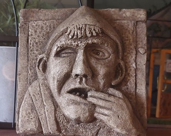 May include: A gray stone sculpture of a man's face with a hand covering his mouth. The sculpture is set in a rectangular frame and has a rough, textured surface.