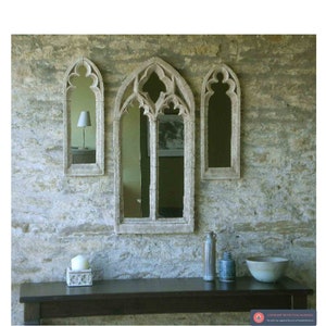 May include: Three arched mirrors with a gothic design are mounted on a stone wall. The mirrors are different sizes and are arranged in a row. A wooden table with a candle and a bowl sits in front of the mirrors.