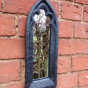 May include: A black gothic-style mirror with a pointed arch top and a decorative frame. The mirror is mounted on a brick wall and reflects a view of a garden.