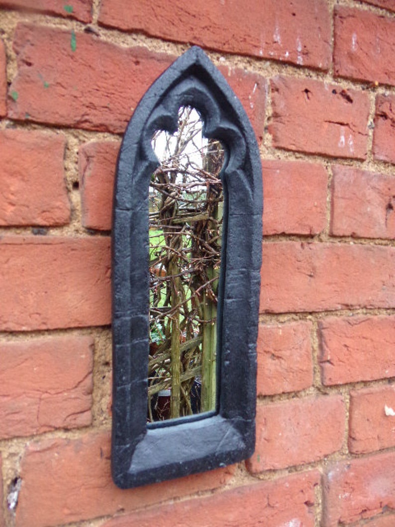 May include: A black gothic-style mirror with a pointed arch top and a decorative frame. The mirror is mounted on a brick wall and reflects a view of a garden.