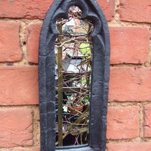 May include: Black metal framed mirror with a gothic arch design. The mirror reflects a view of a garden with plants and vines.