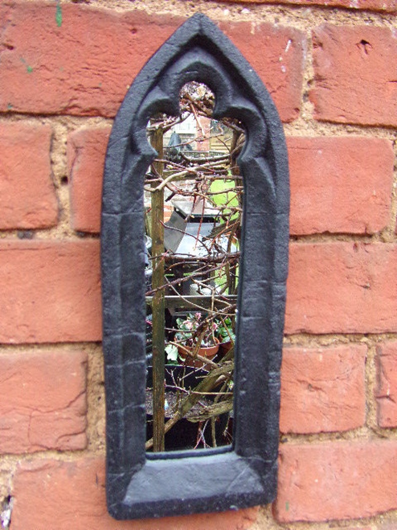 May include: Black metal framed mirror with a gothic arch design. The mirror reflects a view of a garden with plants and vines.