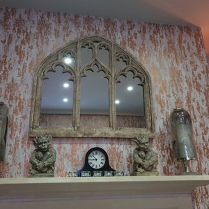 May include: A decorative fireplace mantel with a large arched mirror, two gargoyle figurines, and two glass cloche candle holders. The wall behind the mantel is painted in a distressed copper and white finish.