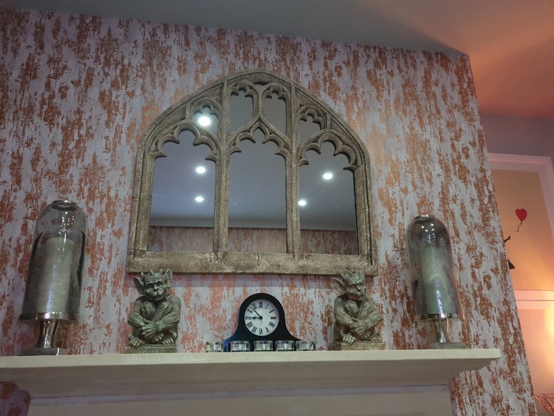 May include: A decorative fireplace mantel with a large arched mirror, two gargoyle figurines, and two glass cloche candle holders. The wall behind the mantel is painted in a distressed copper and white finish.