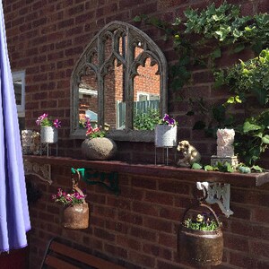 May include: A brick wall with a decorative shelf and a mirror with an arched top. The shelf is decorated with potted plants, a stone, and a small statue. There is a purple umbrella to the left of the shelf.