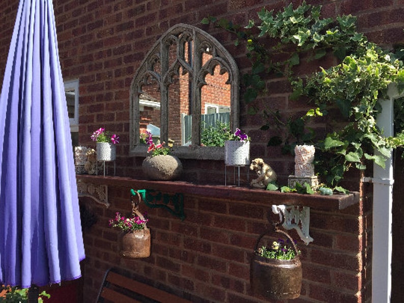 May include: A brick wall with a decorative shelf and a mirror with an arched top. The shelf is decorated with potted plants, a stone, and a small statue. There is a purple umbrella to the left of the shelf.