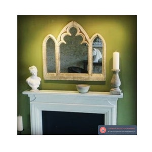 May include: A white, arched, decorative mirror with a gothic design hangs above a white fireplace mantel. The mirror is set against a green wall. A white bust of a woman sits on the mantel, and a small, white bowl sits to the right of the bust. A candle sits on a gray stone candle holder to the right of the bowl.