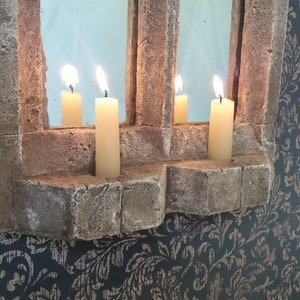 May include: A stone window frame with a mirror inside and four lit candles. The candles are in the stone frame and are lit with a warm yellow glow.