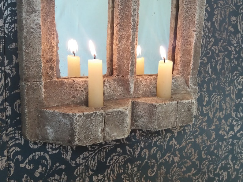 May include: A stone window frame with a mirror inside and four lit candles. The candles are in the stone frame and are lit with a warm yellow glow.