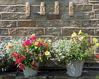 May include: Three white planter pots with a variety of colorful flowers, including red, yellow, and white, are displayed on a stone wall with four decorative stone figures.