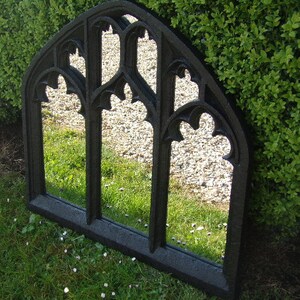 May include: A black metal arched mirror with a gothic design. The mirror is reflecting a green lawn and a stone path.