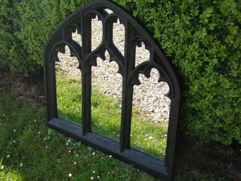 May include: A black metal arched mirror with a gothic design. The mirror is reflecting a green lawn and a stone path.