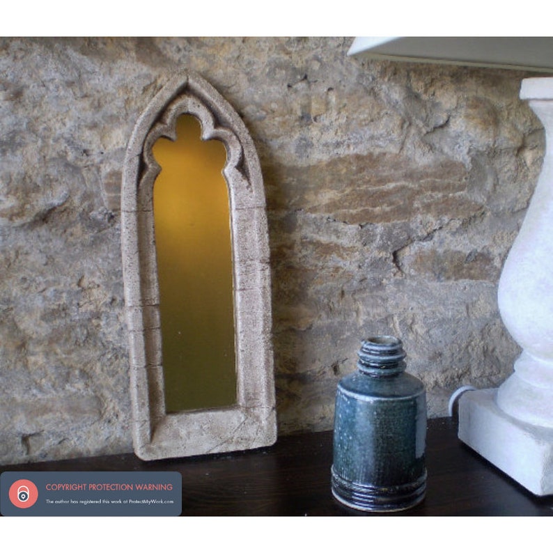 May include: A small, decorative mirror with a stone-like frame in the shape of a gothic arch. The mirror is reflecting a warm, yellow light. The mirror is sitting on a dark wooden surface.