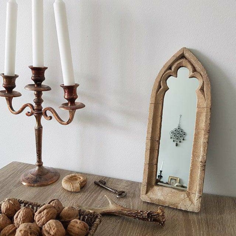 May include: A decorative arched mirror with a light brown finish and a beveled edge. The mirror is framed with a carved design that resembles a stone archway.