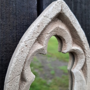 May include: A white, ornate, arched mirror frame with a textured surface. The mirror is reflecting a blurred green background.