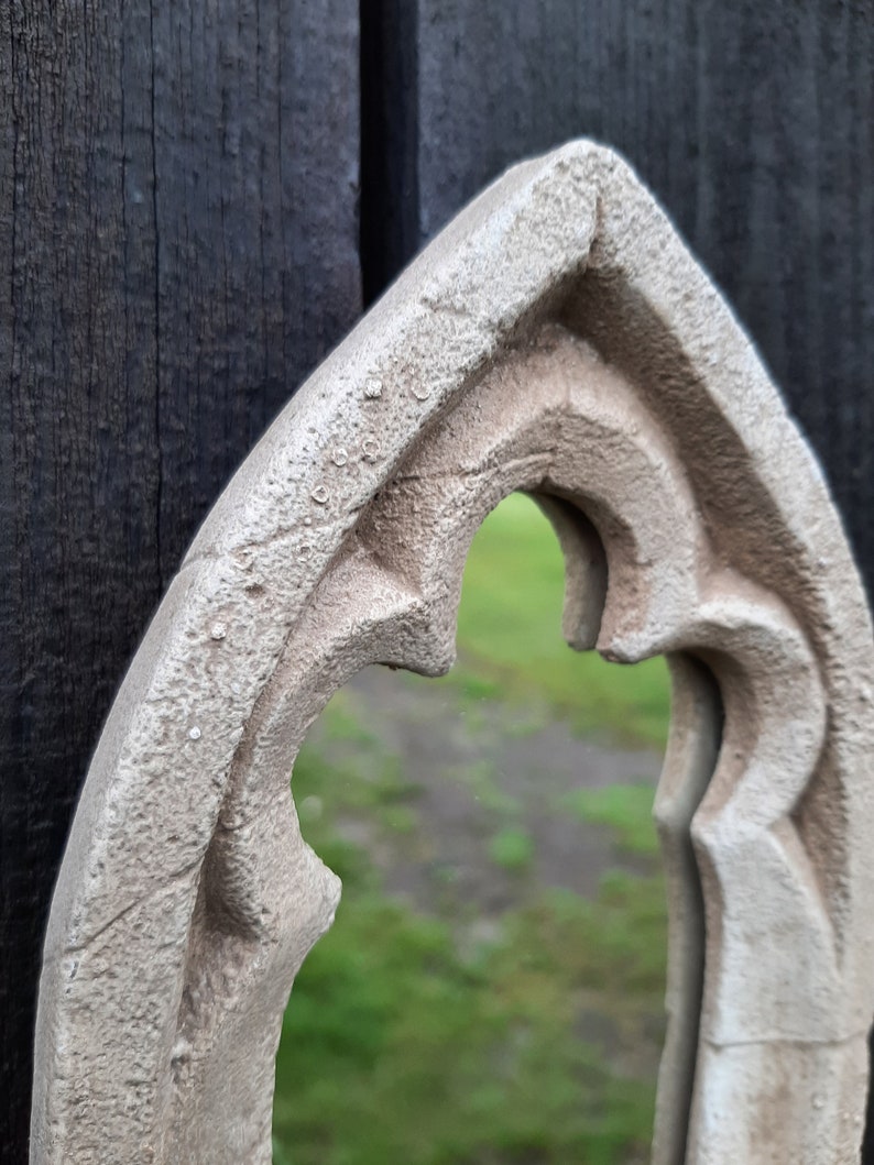 May include: A white, ornate, arched mirror frame with a textured surface. The mirror is reflecting a blurred green background.