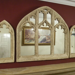 May include: A set of three arched mirrors with ornate frames, each with a different reflection. The center mirror reflects two framed paintings, while the side mirrors reflect a room with a white door. The mirrors are made of a light beige material and have a distressed finish.