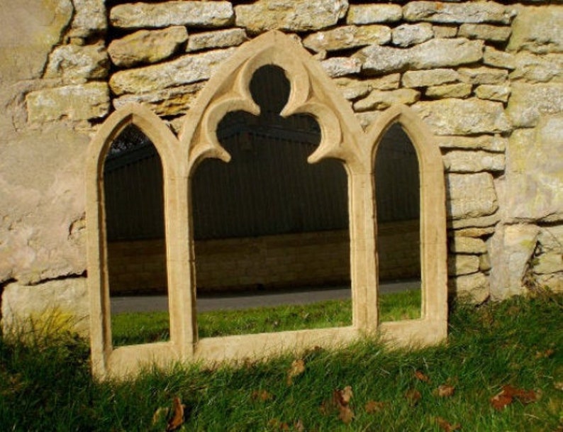 May include: A decorative stone window mirror with a gothic arch design. The mirror has three panels and is made of a light brown stone.