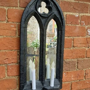 May include: Black gothic style mirror with a pointed arch top and two candle holders. The mirror is reflecting a view of a brick building and trees.