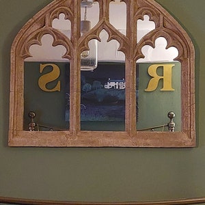 May include: A decorative arched mirror with a stone-like finish, featuring three panels with intricate carvings. The mirror reflects a room with a green wall and a bed with a yellow blanket and white pillow.