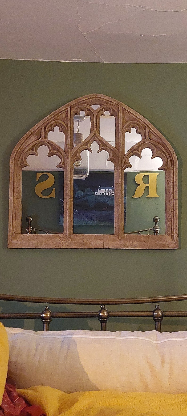 May include: A decorative arched mirror with a stone-like finish, featuring three panels with intricate carvings. The mirror reflects a room with a green wall and a bed with a yellow blanket and white pillow.